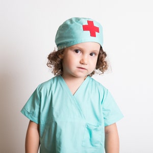 May include: A young child dressed as a doctor, wearing a light blue scrub top and a matching light blue surgical cap with a red cross.