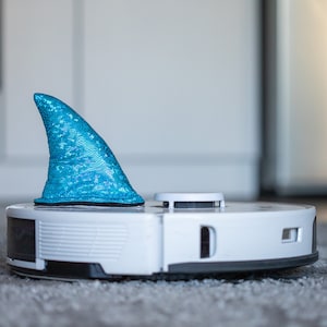 May include: A white robotic vacuum cleaner with a blue sparkly shark fin on top.