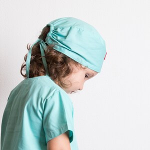 May include: A young person wearing a light blue surgical scrub top and matching hat. The hat has a red accent.