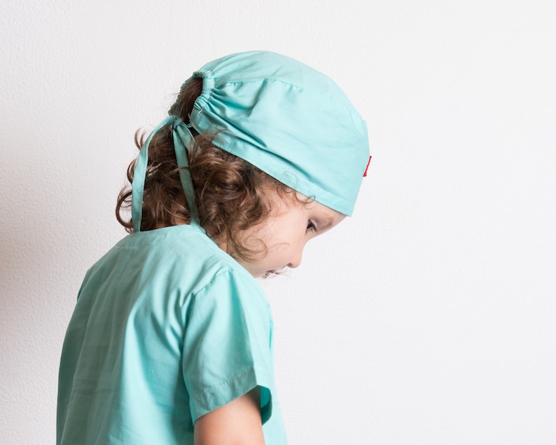 May include: A young person wearing a light blue surgical scrub top and matching hat. The hat has a red accent.