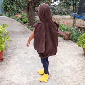 Puede incluir: Un disfraz con capucha de color marr&oacute;n que presenta paneles en forma de alas en la espalda, combinado con leggings azul oscuro y cubrepi&eacute;s de color amarillo brillante. La prenda tiene una textura suave y un dobladillo el&aacute;stico.