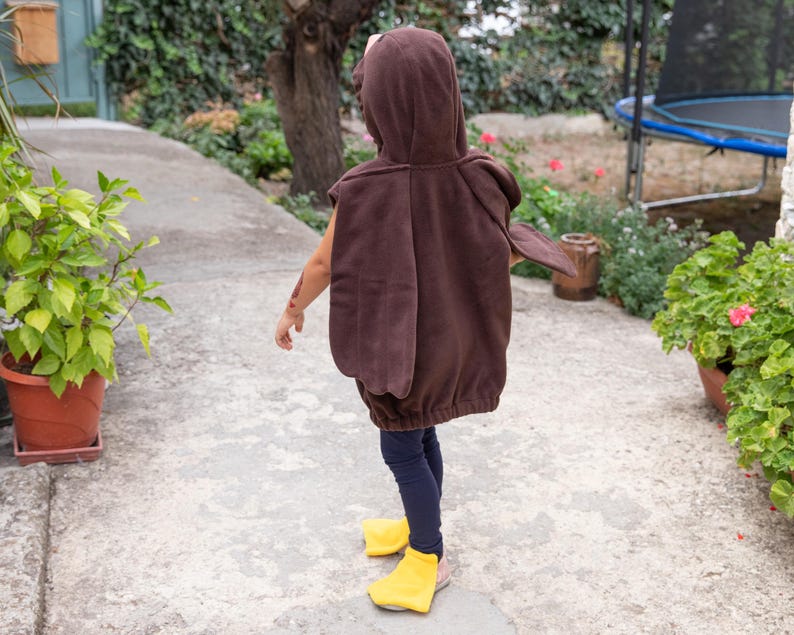 Puede incluir: Un disfraz con capucha de color marr&oacute;n que presenta paneles en forma de alas en la espalda, combinado con leggings azul oscuro y cubrepi&eacute;s de color amarillo brillante. La prenda tiene una textura suave y un dobladillo el&aacute;stico.