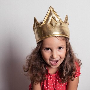 May include: A young person wearing a gold crown and a red polka dot shirt. The person is smiling and looking at the camera.