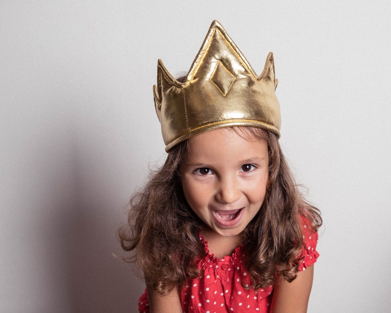 May include: A young person wearing a gold crown and a red polka dot shirt. The person is smiling and looking at the camera.