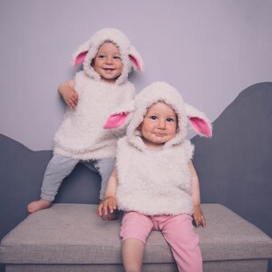 May include: Two toddlers wearing white fluffy sheep costumes with pink ears. They are sitting on a light gray ottoman. The child on the left is standing on the ottoman with one foot on the ground. The child on the right is sitting with their legs crossed.