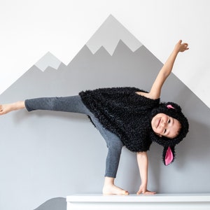 May include: A child dressed in a black sheep costume is balancing on one hand on a white surface. The child is smiling and has their leg raised in the air. The background is a light gray wall with a painted gray mountain design.
