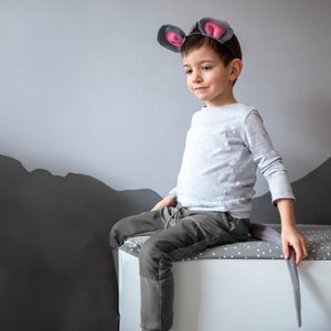May include: A young child wearing a gray mouse costume with pink ears and a tail sits on a white bench with gray polka dots. The child is looking down and smiling.