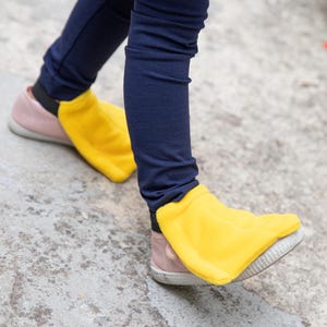 Puede incluir: Un par de cubrezapatos de forro polar amarillo brillante sobre zapatillas de deporte rosa claro, con una banda el&aacute;stica negra en el tal&oacute;n para asegurar la tela sobre el calzado, combinados con unos leggings azul marino oscuro.