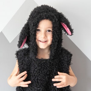 May include: A child wearing a black fluffy sheep costume with pink ears. The costume has a hood and a sleeveless vest.