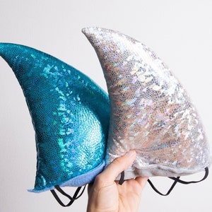 Puede incluir: Dos diademas de cola de sirena, una azul y otra plateada, con un acabado brillante. Las diademas est&aacute;n hechas de tela y tienen cintas el&aacute;sticas.