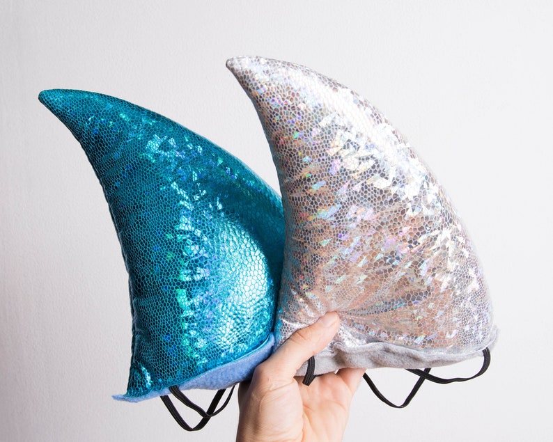 Puede incluir: Dos diademas de cola de sirena, una azul y otra plateada, con un acabado brillante. Las diademas est&aacute;n hechas de tela y tienen cintas el&aacute;sticas.
