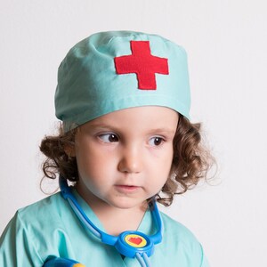 May include: A young child dressed as a doctor wearing a light blue scrub top and matching hat with a red cross. The child is wearing a blue stethoscope around their neck.