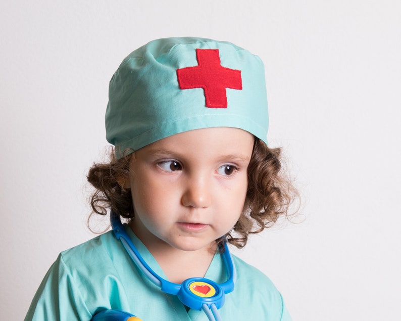 May include: A young child dressed as a doctor wearing a light blue scrub top and matching hat with a red cross. The child is wearing a blue stethoscope around their neck.