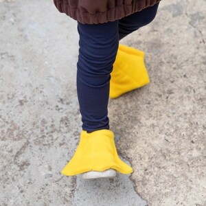 Puede incluir: Un par de fundas de tela de color amarillo brillante sobre zapatos blancos, combinadas con leggings azul marino y una parte superior marr&oacute;n. Las fundas amarillas tienen forma de patas de pato, con un dise&ntilde;o ancho y plano sobre el calzado.