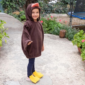 Puede incluir: Un poncho marr&oacute;n con capucha que presenta un dise&ntilde;o de cabeza de pato con un pico naranja, combinado con leggings azul oscuro y cubrepi&eacute;s de color amarillo brillante en forma de patas de pato. El poncho tiene mangas largas y dobladillo el&aacute;stico.