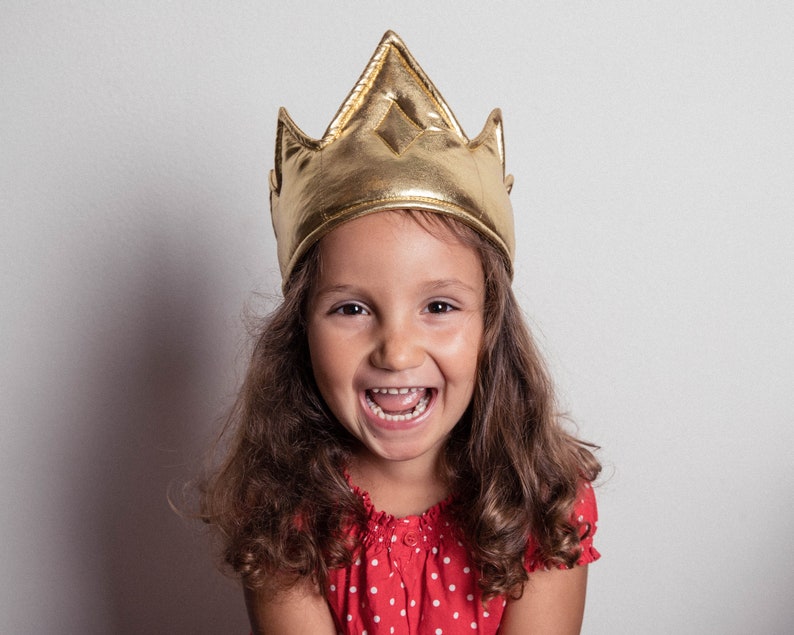 May include: A young girl wearing a gold crown smiles with her mouth open. She has long brown hair and is wearing a red and white polka dot dress.