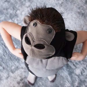 Könnte beinhalten: Ein Gorilla-Kostüm für Kinder, mit einem schwarz-grauen Design und einer Kunstfellkapuze. Das Kostüm hat ein graues Gesicht mit Knopfaugen und einen schwarzen Körper. Das Kind trägt weiße Schuhe.