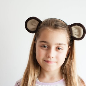 Può includere: Una ragazza giovane con lunghi capelli biondi che indossa una fascia marrone con orecchie di orso pelose. Sta guardando la telecamera con un leggero sorriso.