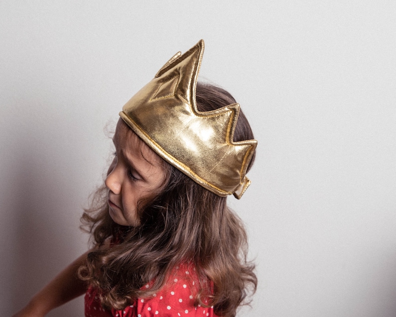 May include: A young person wearing a gold crown with a pointed top. The crown is made of fabric and has a shiny gold finish.