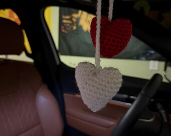 May include: Two crocheted hearts hanging from the rearview mirror of a car. The top heart is red and the bottom heart is white.
