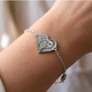 May include: A silver bracelet with a heart-shaped pendant, engraved with a portrait of a couple. The bracelet is made of a fine chain and is worn on a wrist. The pendant's design is a detailed portrait.