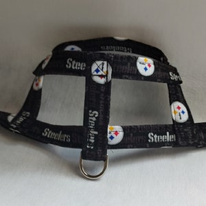 May include: Black dog harness featuring the Pittsburgh Steelers logo in white, yellow, and red. The harness has a black buckle and a metal D-ring for leash attachment. The word "Steelers" is printed on the straps.