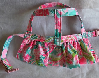 May include: A dog harness with a pink, green, and turquoise floral pattern. The harness has a skirt-like design and a pink plastic buckle. The harness has a metal ring for leash attachment.