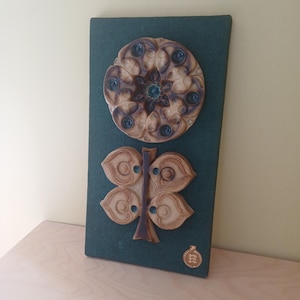 May include: A rectangular dark green fabric-covered wall plaque featuring two ceramic relief sculptures. The top piece is a circular floral design, and the bottom piece is a four-leaf clover shape. A small gold-colored circular sticker is at the bottom.