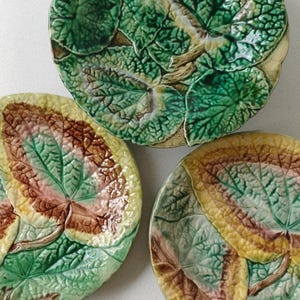 May include: Three ceramic plates with a leaf design. The plates feature a raised leaf pattern in shades of green, brown, and yellow. The plates have a rustic, vintage aesthetic.