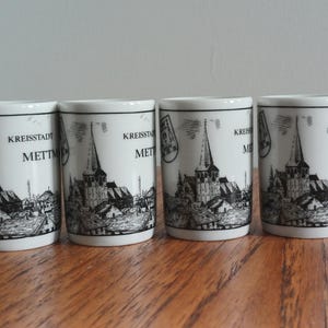 Pode incluir: Quatro copos de shot de cerâmica branca com uma ilustração a preto e branco de uma igreja e o texto "Kreisstadt Mett". Os copos são cilíndricos e têm uma borda preta.