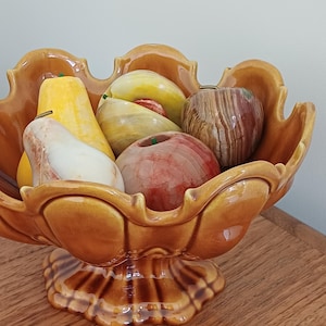 Pode incluir: Uma fruteira de cerâmica em tom âmbar quente, cheia de frutas decorativas de pedra. A tigela tem uma borda ondulada e uma base de pedestal. As frutas incluem peras, maçãs e outras frutas de caroço em várias cores e texturas.