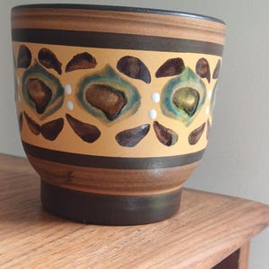 May include: A ceramic planter with a brown and tan color scheme. The planter features a repeating pattern of teal and brown shapes with white dots on a tan background. The top and bottom rims are dark brown.