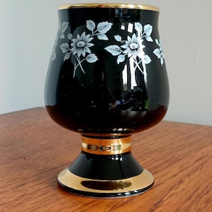 Pode incluir: Um vaso de vidro preto com borda e base douradas. Desenhos florais brancos decoram o corpo do vaso. O vaso tem formato bulboso e assenta sobre uma base curta em forma de pedestal. O vaso tem aproximadamente 15 cm de altura.