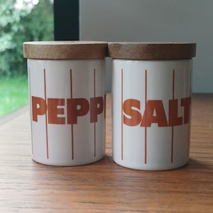May include: Two white ceramic salt and pepper pots with wooden lids. The words "PEPPER" and "SALT" are printed in orange block letters on the front of each pot. Vertical orange lines are printed on the white background.