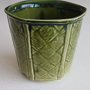 Peut inclure: Un pot de plante en céramique verte avec un motif de panier tissé texturé. Le pot a une forme carrée avec des coins arrondis et un bord vert foncé. L'extérieur présente des accents verticaux en forme de bambou.