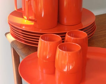May include: A set of orange-colored dinnerware, including mugs, plates, and tumblers. The mugs have handles, and the plates are stacked. The tumblers and plates are on a large oval tray. The items are arranged on a wooden table.