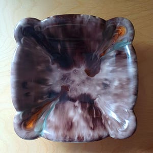 Könnte beinhalten: Ein quadratischer Aschenbecher aus Keramik mit einem Wirbelmuster in Braun-, Weiß- und Blautönen. Der Aschenbecher hat vier abgerundete Ecken und einen leicht erhöhten Rand.