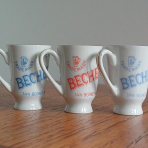 Pode incluir: Três canecas de cerâmica branca com alça, cada uma com o texto BECHEROVKA, um pequeno ícone de caneca e as palavras TRADEMARK e JAN BECHER. Duas canecas têm letras azuis, enquanto a caneca central apresenta o mesmo texto e ícones em vermelho.