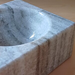 Pode incluir: Uma tigela de m&aacute;rmore branco com um interior liso e arredondado. A tigela tem uma forma quadrada e um padr&atilde;o veado cinza.