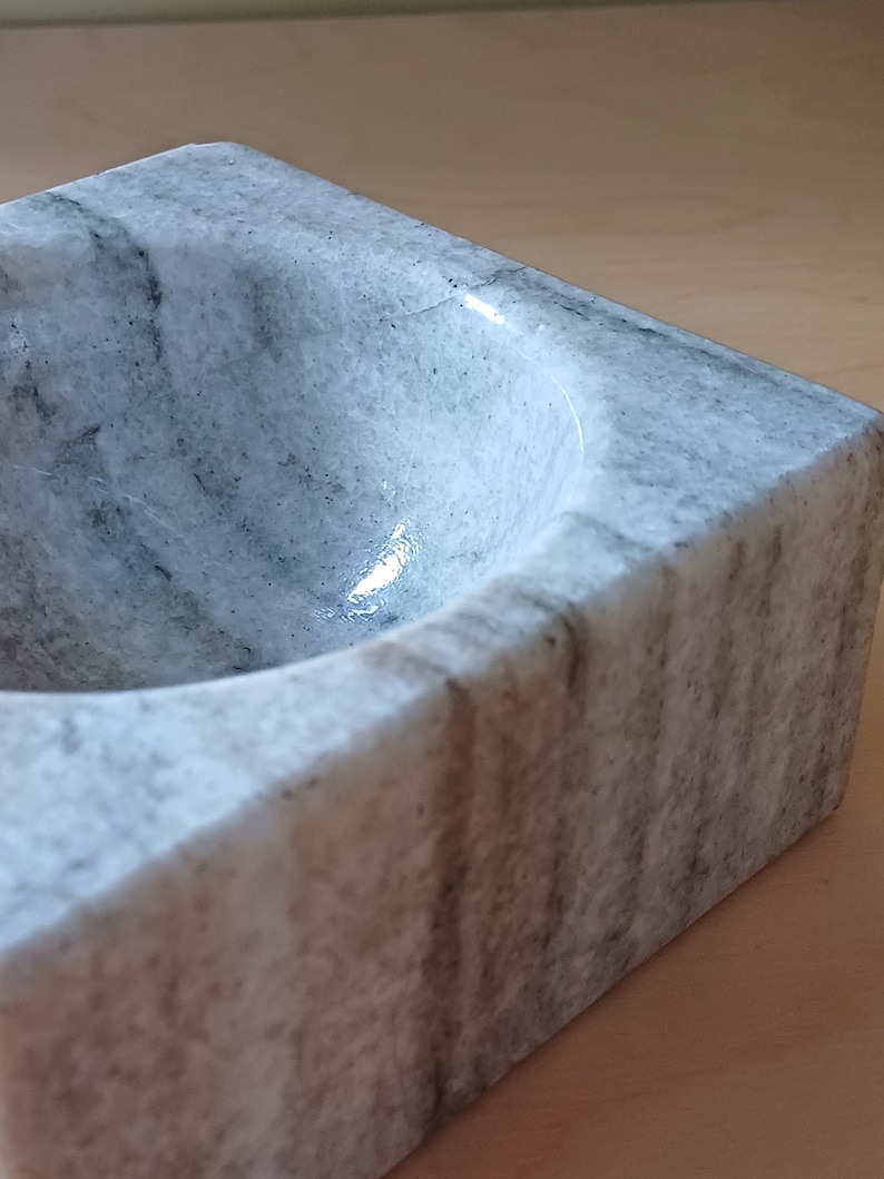 Pode incluir: Uma tigela de m&aacute;rmore branco com um interior liso e arredondado. A tigela tem uma forma quadrada e um padr&atilde;o veado cinza.