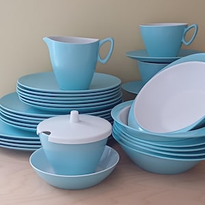May include: A full set of light blue tableware, including plates, bowls, cups, saucers, a creamer, and a lidded sugar bowl. The plates and bowls are stacked, showing the full set. The interior of the bowls and creamer are white.