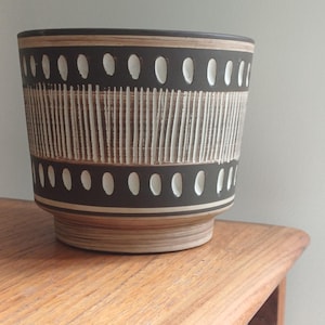 May include: A cylindrical, brown ceramic planter with a textured design. The planter features alternating bands of vertical lines and oval shapes in white against a dark brown background. The planter sits on a light brown base.