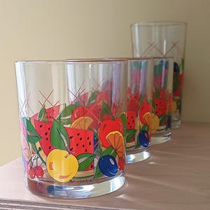 Könnte beinhalten: Set aus vier transparenten Trinkgläsern mit einem farbenfrohen Fruchtdesign. Die Gläser zeigen Illustrationen von Wassermelone, Orangen, Kirschen und Pflaumen. Der Text "By Cer E" ist auf der Unterseite des Glases aufgedruckt.