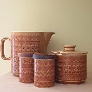 May include: A collection of brown ceramic kitchen items. The set comprises a jug, two lidded canisters, and a lidded jar. Each piece has a repeating floral design and a warm, earthy colour, ideal for a retro kitchen setting.