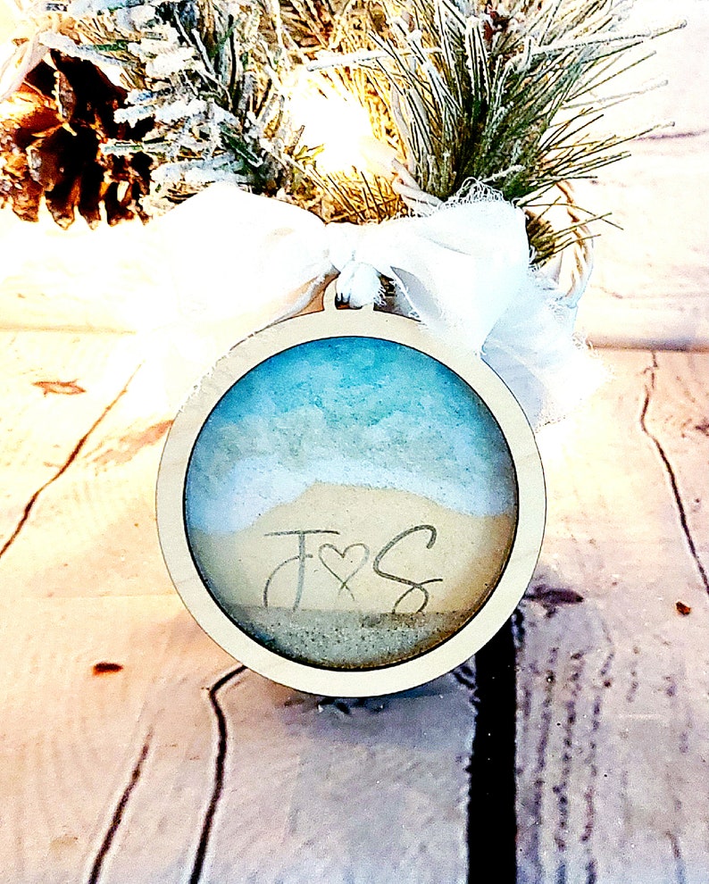 May include: A round wooden ornament with a blue and white beach scene inside. The ornament has the initials "J & S" written in the sand.
