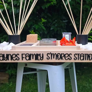 May include: A wooden s'mores station with the text "Haynes Family S'mores Station" on the front. The station includes compartments for graham crackers, marshmallows, chocolate, and skewers. The station is set on a white table.