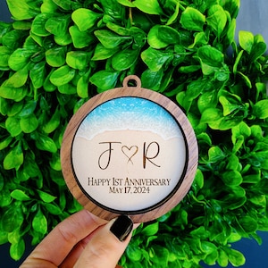 May include: A round wooden ornament with a beach scene and the initials "J & R" engraved on it. The text "Happy 1st Anniversary May 17, 2024" is also engraved on the ornament.