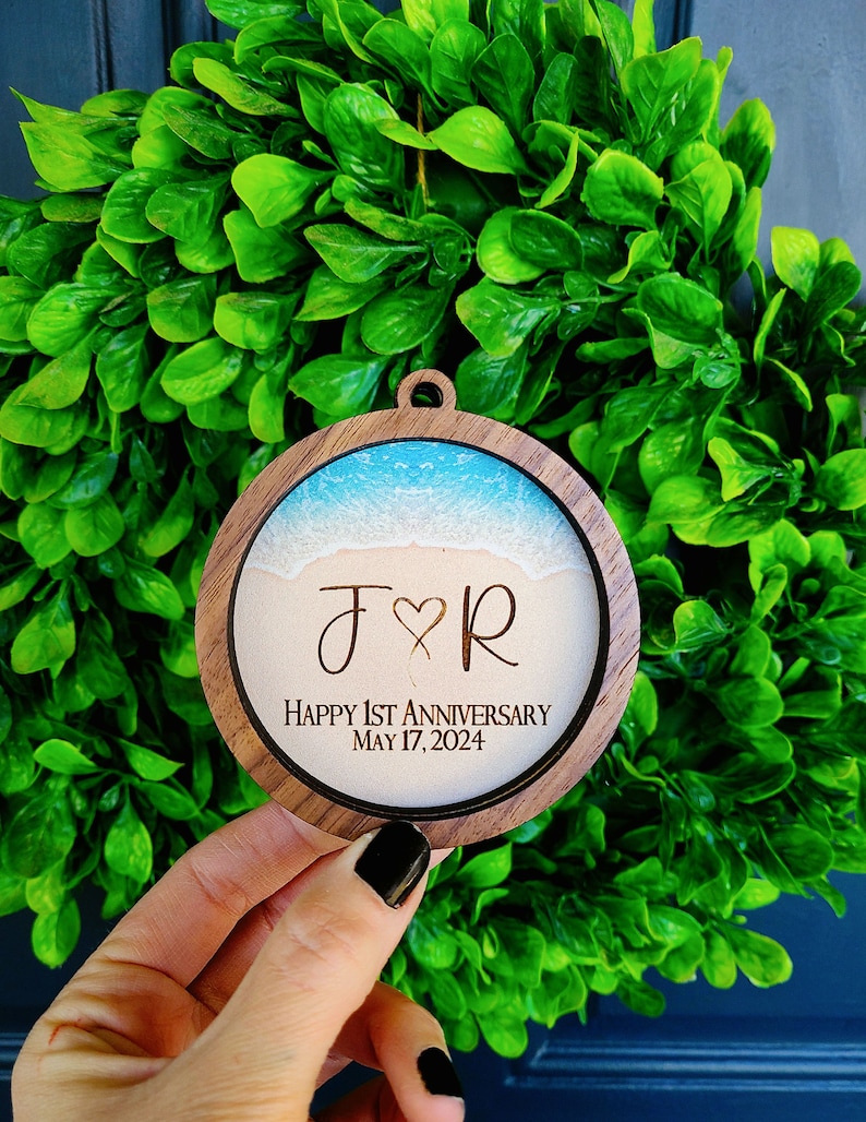 May include: A round wooden ornament with a beach scene and the initials "J & R" engraved on it. The text "Happy 1st Anniversary May 17, 2024" is also engraved on the ornament.