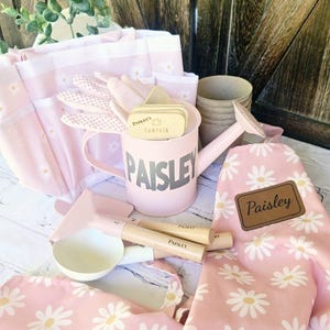 May include: A pink gardening set with daisy print fabric and tools. The set includes a watering can with the name "Paisley", gloves, a tote bag, and a trowel. The tools have wooden handles. The set is designed for gardening.