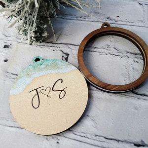 May include: A wooden ornament with a light blue and white watercolor design resembling a beach scene. The ornament has the initials "J & S" with a heart symbol in between. The ornament is attached to a wooden hoop.